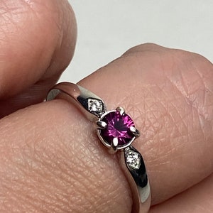 Op de afbeelding: Een zilveren ring met een ronde, donkerroze edelsteen omgeven door kleinere, heldere edelstenen.