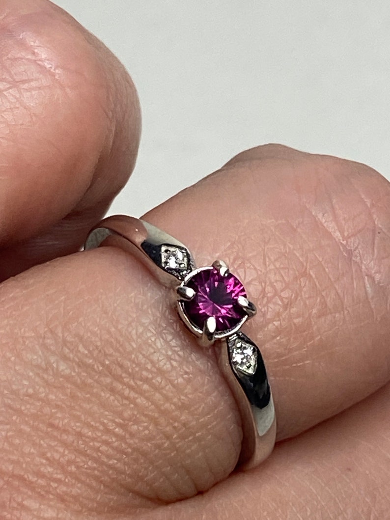 Op de afbeelding: Een zilveren ring met een ronde, donkerroze edelsteen omgeven door kleinere, heldere edelstenen.