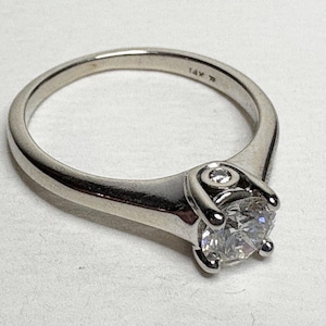 May include: A silver ring with a large, clear, round-cut gemstone. The ring has a simple, elegant design with a single, smaller accent stone. The band is marked "14K".