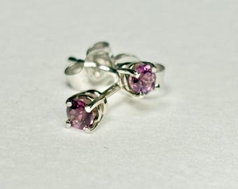 May include: A pair of silver stud earrings with round, light purple gemstones. The earrings are set in a four-prong setting, and the posts and backs are visible. The earrings are on a white surface.