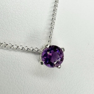 May include: A silver necklace featuring a round, deep purple gemstone pendant. The gemstone is set in a four-prong setting and is suspended from a delicate chain. The chain and setting appear to be made of silver.