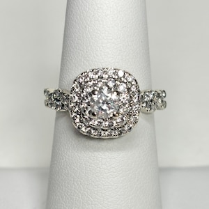 May include: A silver ring with a square halo of small, clear stones surrounding a larger, central clear stone. The band features additional small stones on each side of the halo, creating a sparkling effect. The ring is displayed on a white surface.