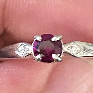 Op de afbeelding: Een zilveren ring met een ronde, donkerrode edelsteen en twee kleinere, heldere edelstenen op de band.