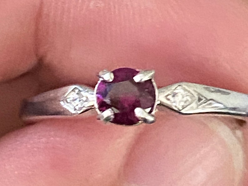 Op de afbeelding: Een zilveren ring met een ronde, donkerrode edelsteen en twee kleinere, heldere edelstenen op de band.