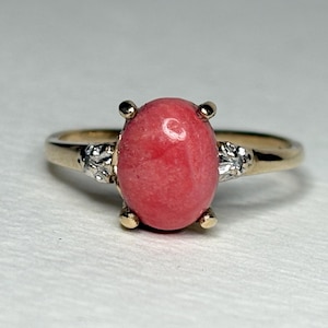 May include: A gold ring with a large, oval, pink gemstone as the centerpiece. The gemstone is held in place by four gold prongs. The band is gold with small, decorative silver accents on either side of the gemstone.