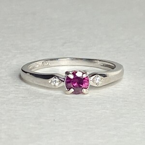 Op de afbeelding: Een zilveren ring met een roze edelsteen en twee kleinere witte edelstenen.