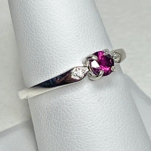 Op de afbeelding: Een zilveren ring met een paarse edelsteen en twee kleinere heldere edelstenen. De ring ligt op een witte displaystandaard.