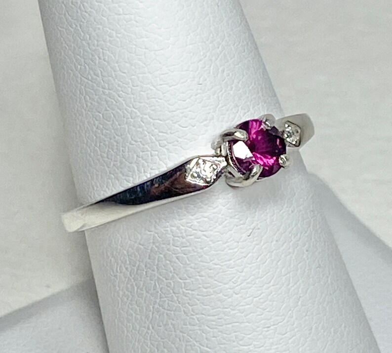 Op de afbeelding: Een zilveren ring met een paarse edelsteen en twee kleinere heldere edelstenen. De ring ligt op een witte displaystandaard.