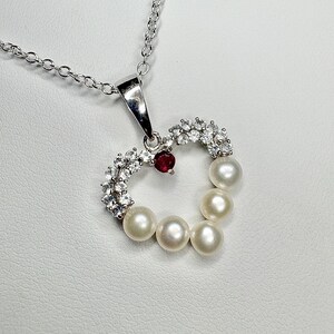 May include: A silver necklace with a heart-shaped pendant. The pendant features a row of white pearls, clear crystals, and a small red gemstone. The necklace chain is delicate and silver.