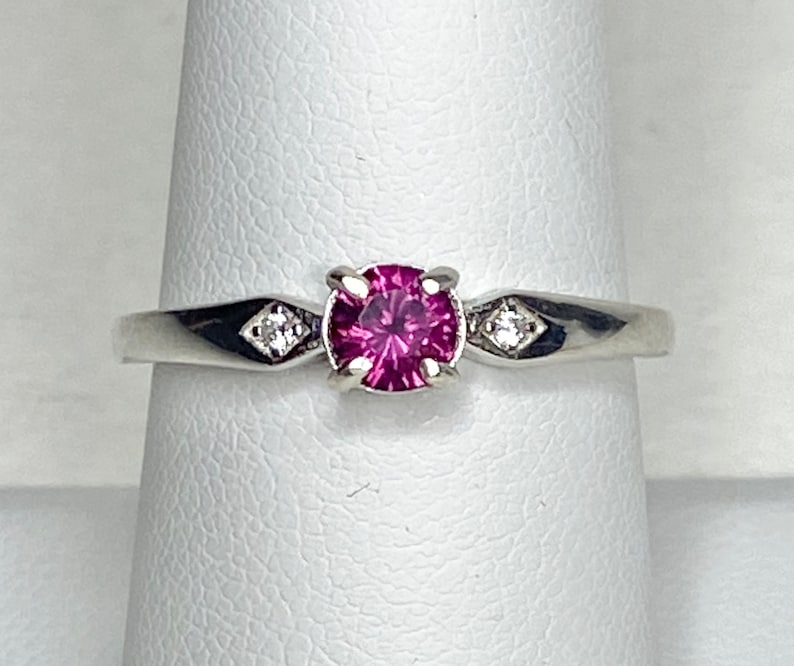 Op de afbeelding: Een zilveren ring met een roze edelsteen en twee kleinere diamanten aan weerszijden.
