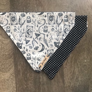 May include: A dog bandana with a beige background and a dark gray tarot card design. The design includes moons, stars, and other celestial elements. The reverse side is black with white polka dots. A small leather tag reads "JoyStar".