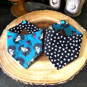 May include: Two triangular pet bandanas with snap closures. One features a teal background with a repeating print of a person with dark hair and red hearts, while the other has a black background with a repeating pattern of white triangles.