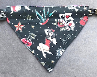May include: A black triangular pet bandana featuring a white polka dot pattern and various colorful tattoo-style graphics including a pin-up figure, a swallow, playing cards with the word LUCKY, cherries, an eight ball, and a star.