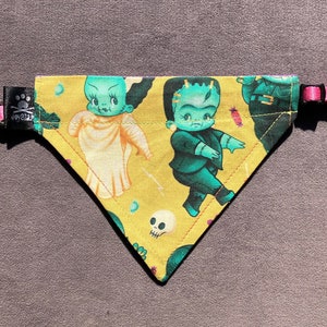 May include: A triangular pet bandana in mustard yellow featuring a repeating pattern of cartoon-style Frankenstein monsters and brides, skulls, and small candies. The bandana includes a black fabric tag with a paw print logo and a pink plastic buckle.