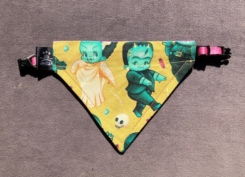 May include: A triangular pet bandana in mustard yellow featuring a repeating pattern of cartoon-style Frankenstein monsters and brides, skulls, and small candies. The bandana includes a black fabric tag with a paw print logo and a pink plastic buckle.