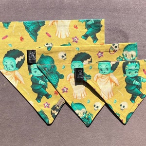 May include: Three yellow fabric dog bandanas with a cupie doll  monster theme. The bandanas feature a repeating pattern of green and blue cartoon monsters with skulls and flowers. The bandanas are arranged in a triangular shape with the largest bandana at the top and the smallest bandana at the bottom.