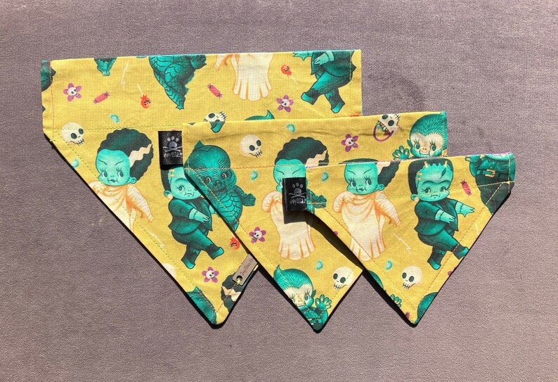 May include: Three yellow fabric dog bandanas with a cupie doll  monster theme. The bandanas feature a repeating pattern of green and blue cartoon monsters with skulls and flowers. The bandanas are arranged in a triangular shape with the largest bandana at the top and the smallest bandana at the bottom.