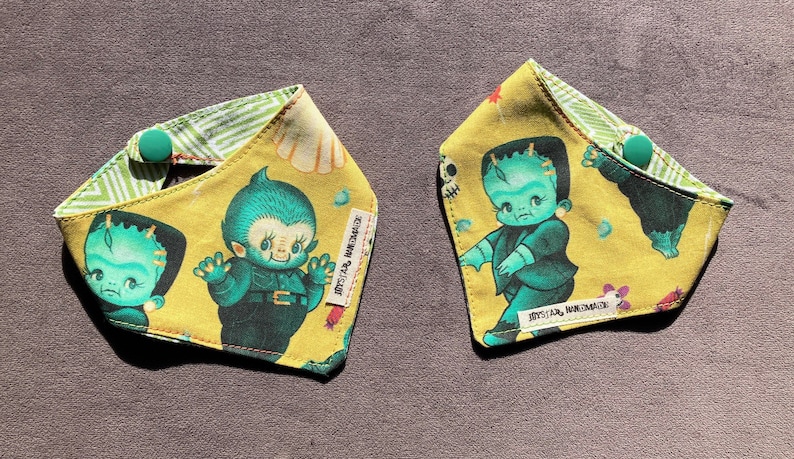 May include: Two triangular fabric bandanas featuring a yellow background with green monster illustrations, including a creature resembling Frankenstein's monster. Each bandana has a green snap button closure and a white tag labeled JAYSSTAR HANDMADE.