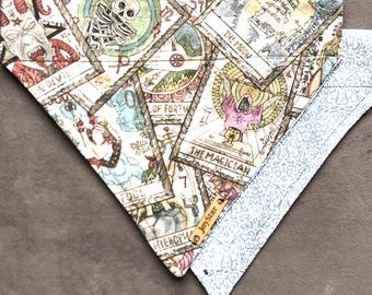 Puede incluir: Un pañuelo triangular para mascotas con un collage de ilustraciones de cartas del tarot, incluyendo El Diablo, La Luna, El Mago, El Loco y El Hierofante. La tela tiene una pequeña etiqueta de cuero marrón con el texto JoyStar. El reverso es azul claro con un patrón de damasco blanco.