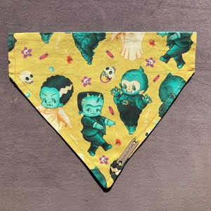 May include: A yellow bandana with a  cupie doll monster theme. The bandana features a variety of green monsters with black eyes and white teeth. The bandana has a white border and is decorated with candy corn and other Halloween-themed designs.