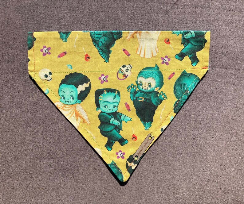 May include: A yellow bandana with a  cupie doll monster theme. The bandana features a variety of green monsters with black eyes and white teeth. The bandana has a white border and is decorated with candy corn and other Halloween-themed designs.