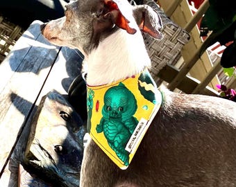May include: A dog bandana featuring a yellow background with a green creature graphic and small red candy illustrations. The fabric accessory has a white tag with black text that reads TOYSTAR HANDMADE and is worn around the neck of a dog.