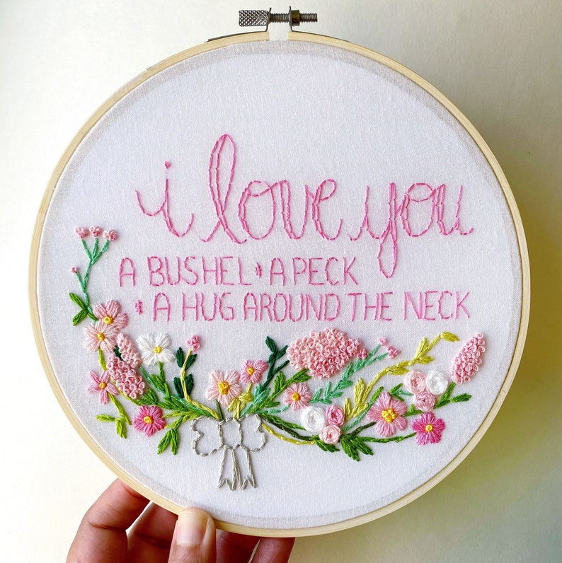 May include: Embroidered hoop art with pink floral design and the text "I love you a bushel, a peck, a hug around the neck."