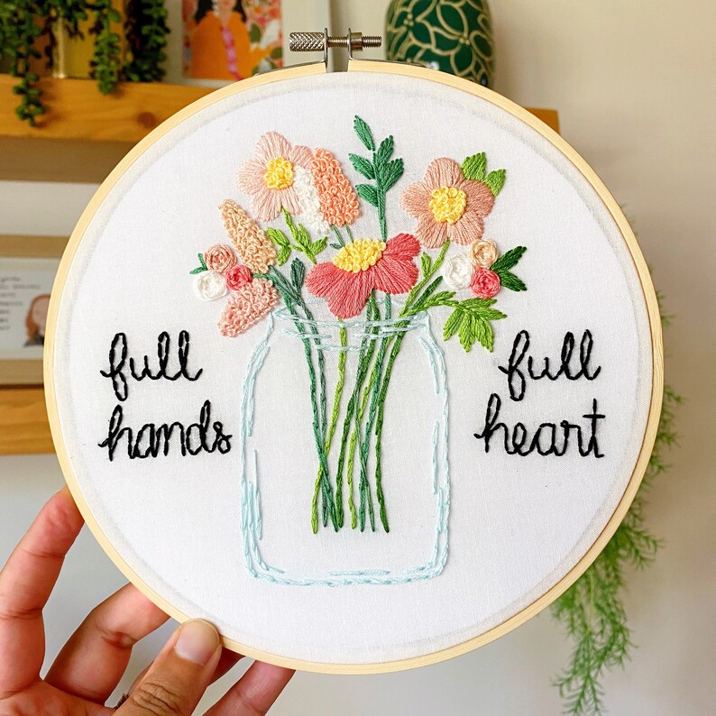 Puede incluir: Arte de bordado en aro con un dise&ntilde;o floral en tonos rosa, verde y blanco. Las flores est&aacute;n dispuestas en un frasco de vidrio con el texto "full hands full heart" bordado debajo.