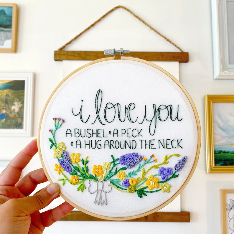 May include: Embroidered hoop art with a white background and green lettering that reads "i love you a bushel, a peck, a hug around the neck". The lettering is surrounded by a wreath of colorful embroidered flowers.