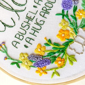 May include: Embroidered hoop art with a floral design featuring purple, yellow, and green flowers. The text "A BUSHEL & A HUG AROUND" is stitched in green thread.