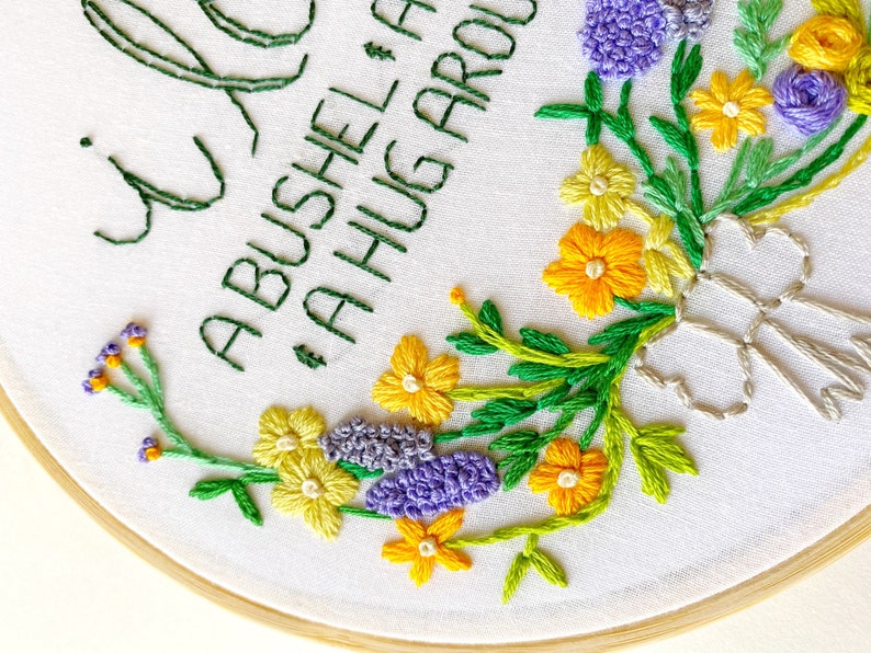 May include: Embroidered hoop art with a floral design featuring purple, yellow, and green flowers. The text "A BUSHEL & A HUG AROUND" is stitched in green thread.