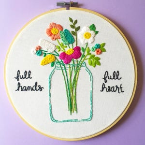 Puede incluir: Arte de bordado en aro con un ramo de flores en un frasco de vidrio. Las flores son rosas, blancas, amarillas y verdes. El texto "full hands full heart" est&aacute; bordado en el aro.