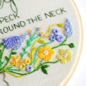 May include: Embroidered hoop art with the text "Give a peck & hug around the neck" and a floral design featuring yellow, green, and purple flowers.