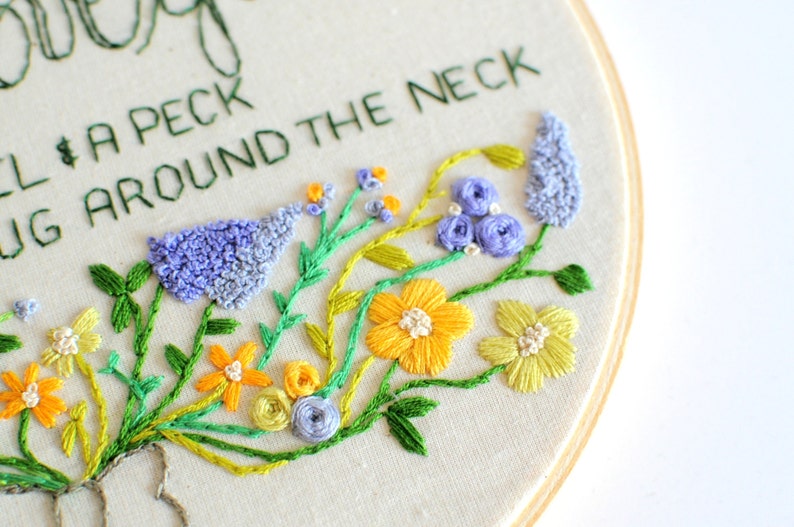 May include: Embroidered hoop art with the text "Give a peck & hug around the neck" and a floral design featuring yellow, green, and purple flowers.