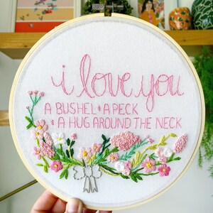 May include: Embroidered hoop art with pink thread on white fabric. The text reads "I love you a bushel and a peck and a hug around the neck."  The text is surrounded by a wreath of pink and green flowers.