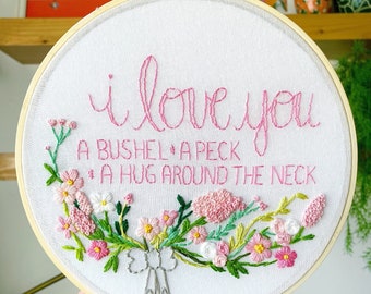 May include: Embroidered hoop art with pink thread on white fabric. The text reads "I love you a bushel and a peck and a hug around the neck."  The text is surrounded by a wreath of pink and green flowers.