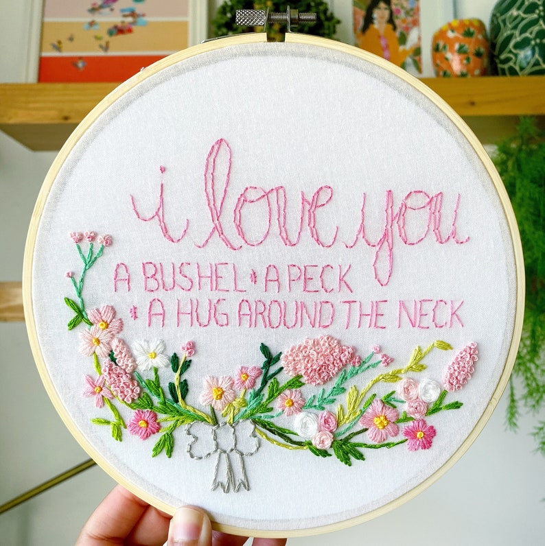May include: Embroidered hoop art with pink thread on white fabric. The text reads "I love you a bushel and a peck and a hug around the neck."  The text is surrounded by a wreath of pink and green flowers.