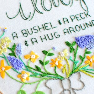 May include: Embroidered hoop art with a floral design and the text "A bushel & a peck & a hug around the neck".
