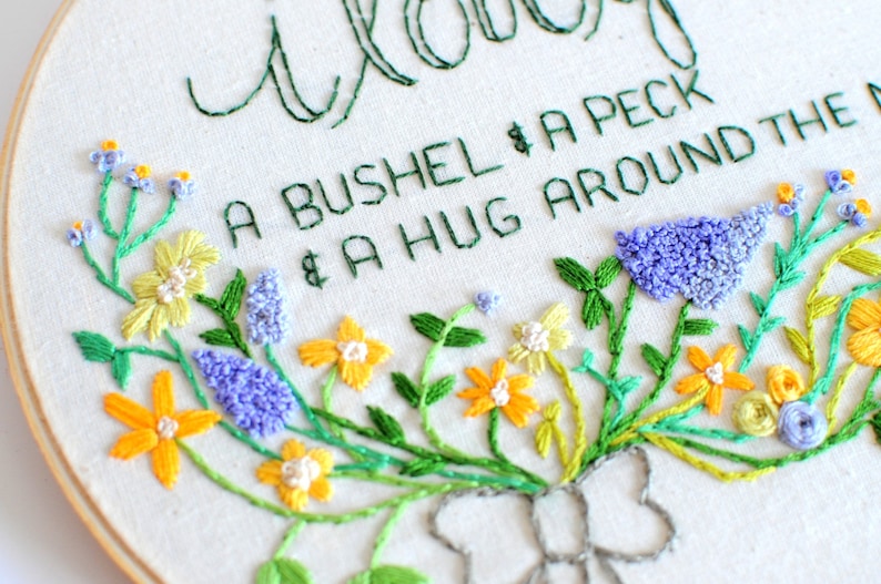 May include: Embroidered hoop art with a floral design and the text "A bushel & a peck & a hug around the neck".