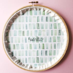 Puede incluir: Un aro de bordado de madera redondo con un respaldo de tela blanca. La tela est&aacute; impresa con un patr&oacute;n de cuadrados de acuarela verde y azul. El texto "handmade by KimArt Designs" est&aacute; impreso en negro en la tela.