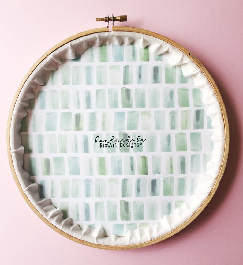 Puede incluir: Un aro de bordado de madera redondo con un respaldo de tela blanca. La tela est&aacute; impresa con un patr&oacute;n de cuadrados de acuarela verde y azul. El texto "handmade by KimArt Designs" est&aacute; impreso en negro en la tela.
