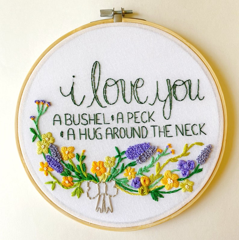 May include: Embroidered hoop art with a white background and green, yellow, and purple floral embroidery. The text "I love you a bushel, a peck, a hug around the neck" is embroidered in green thread.