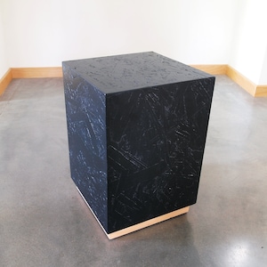 May include: A black square pedestal with a wood base. The pedestal is made of a textured material that resembles wood.