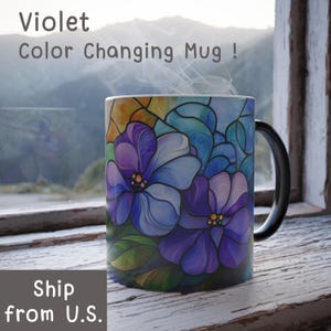 May include: A color-changing mug with a black handle. The mug features a stained glass design of purple and blue flowers. The text "Violet Color Changing Mug!" is visible at the top of the image.