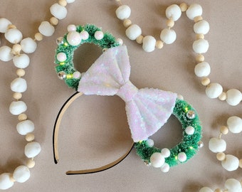 May include: A pair of white and green Minnie Mouse ears with a white bow. The ears are decorated with white beads and green sprigs.
