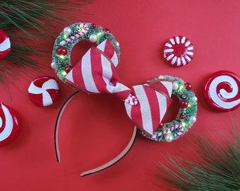 May include: Red and white striped bow with a green and white wreath on each side. The wreath is decorated with candy canes, white beads, and red berries. The bow and wreaths are attached to a black headband.