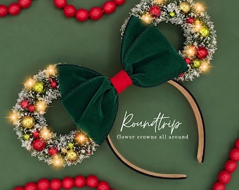 May include: A headband featuring two miniature wreaths decorated with red and gold ornaments, dusted with artificial snow, and illuminated by small lights. A large dark green velvet bow with a red center sits between the wreaths. Text reads Roundtrip flower crowns all around.