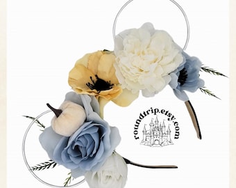 May include: A pair of white mouse ears with a floral arrangement. The flowers are blue, white, yellow, and cream. There is a small white pumpkin on the left ear. The text "roundtrip.etsy.com" is visible on the headband.