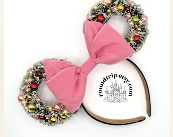 May include: Pink bow headband with two green and white Christmas wreaths decorated with red and gold ornaments. The headband has a black band with a white castle logo and the text "roundtrip.etsy.com".