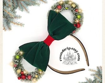 May include: Green velvet bow with a red ribbon, topped with two Christmas wreaths decorated with red and green ornaments, white snow, and gold glitter. The wreaths are attached to a brown headband. The text "roundtrip.etsy.com" is printed on the headband.